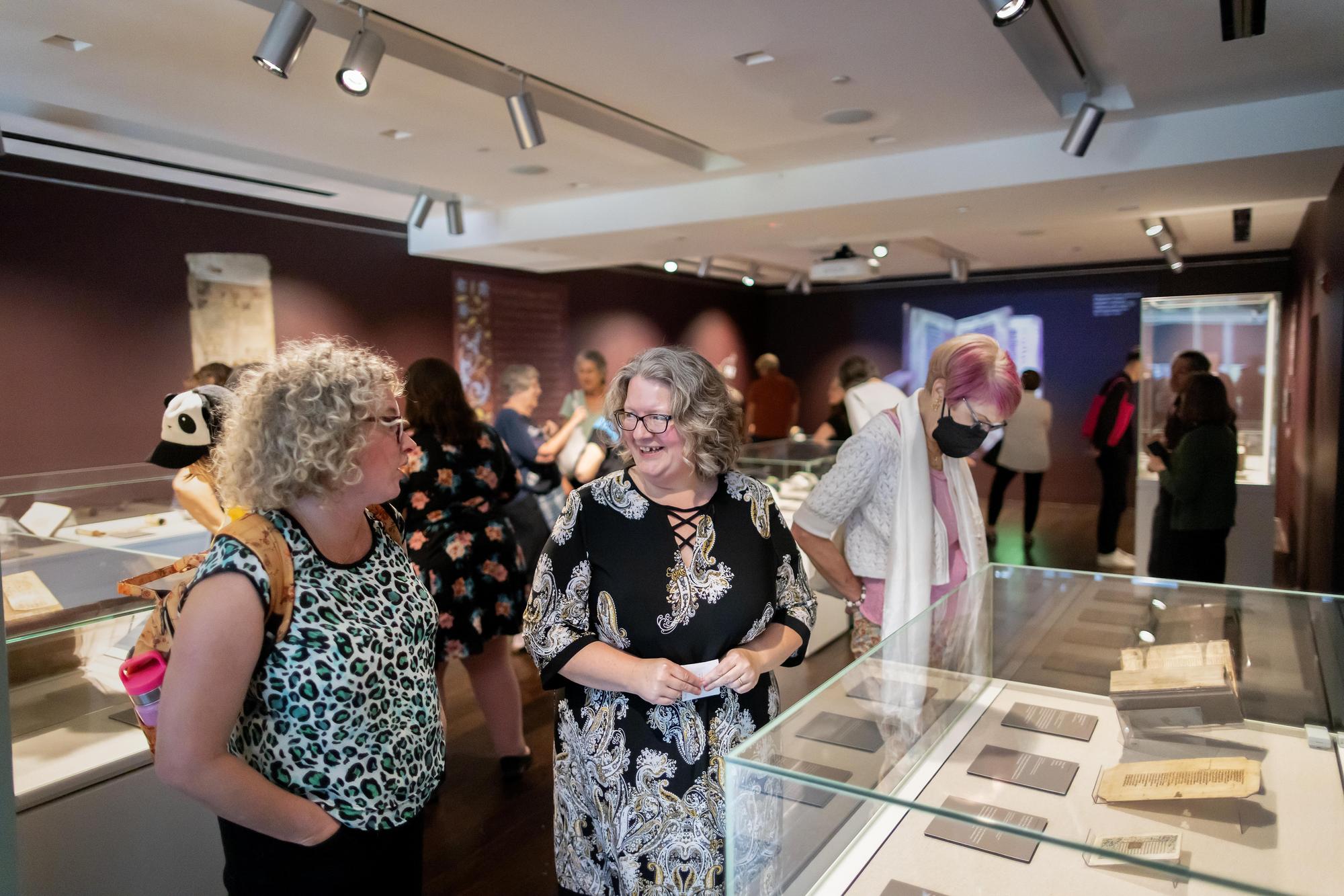 Dot Porter standing and talking with another person in front of a glass case that has books in it in a gallery setting. 