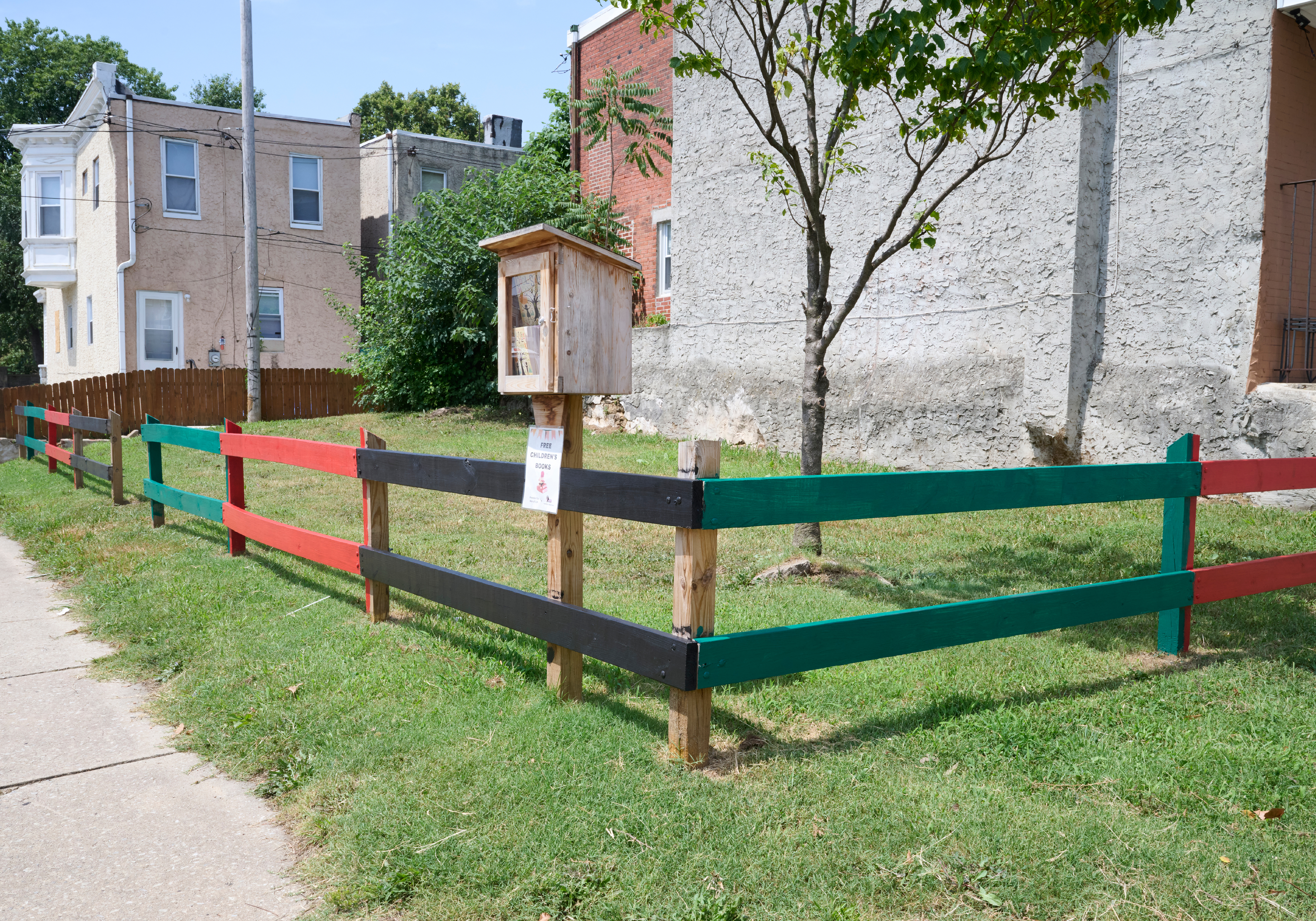 A grassy corner lot adjacent to rowhouses is surrounded by a painted wooden fence, with a little free library kiosk in front.