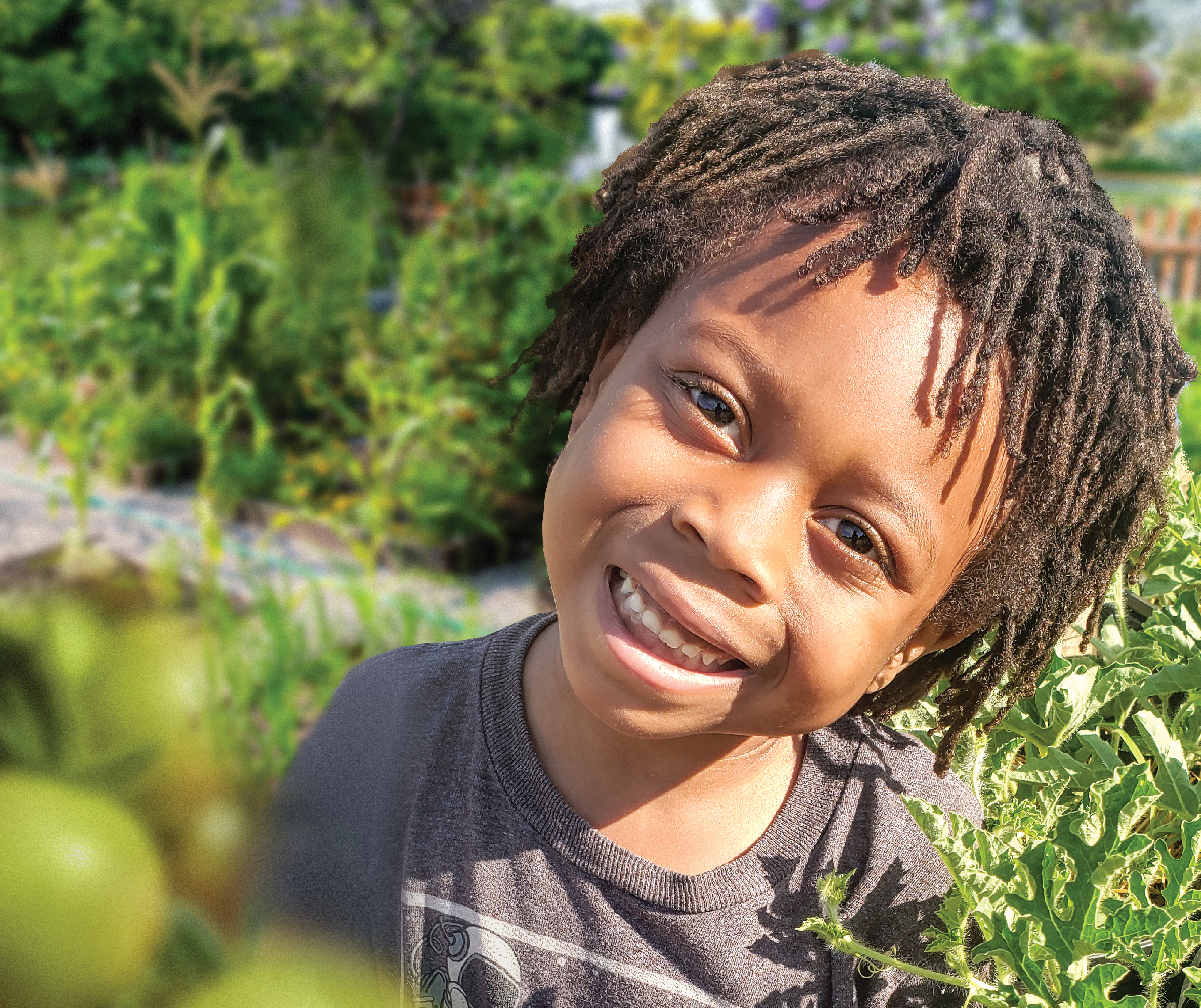 A young child smiles at the camera, surrounded by green growing plants