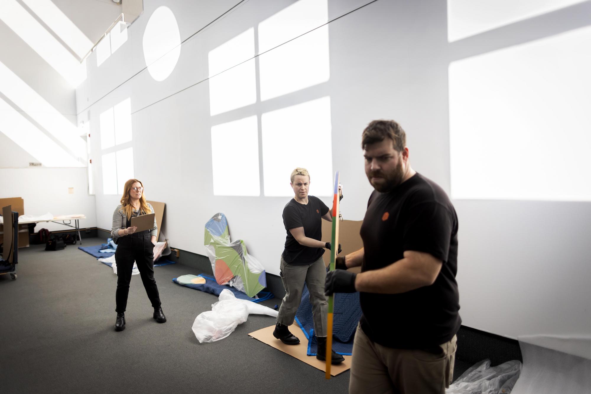 Lynn Dolby standing in a high-ceilinged space observing workers holding an artwork