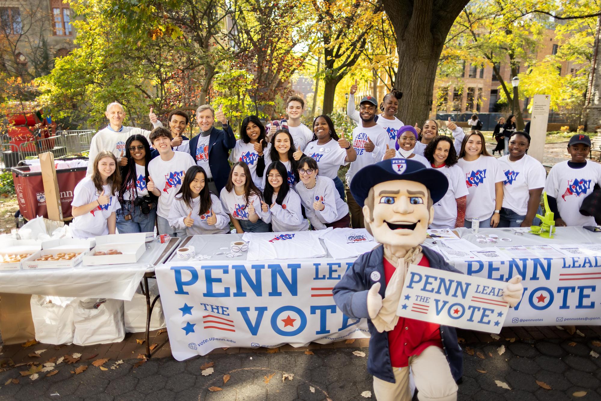 Interim President J. Larry Jameson and members of Penn Leads the Vote pose behind a table with their thumbs up to promote voting. The Quaker mascot is in front on one knee.