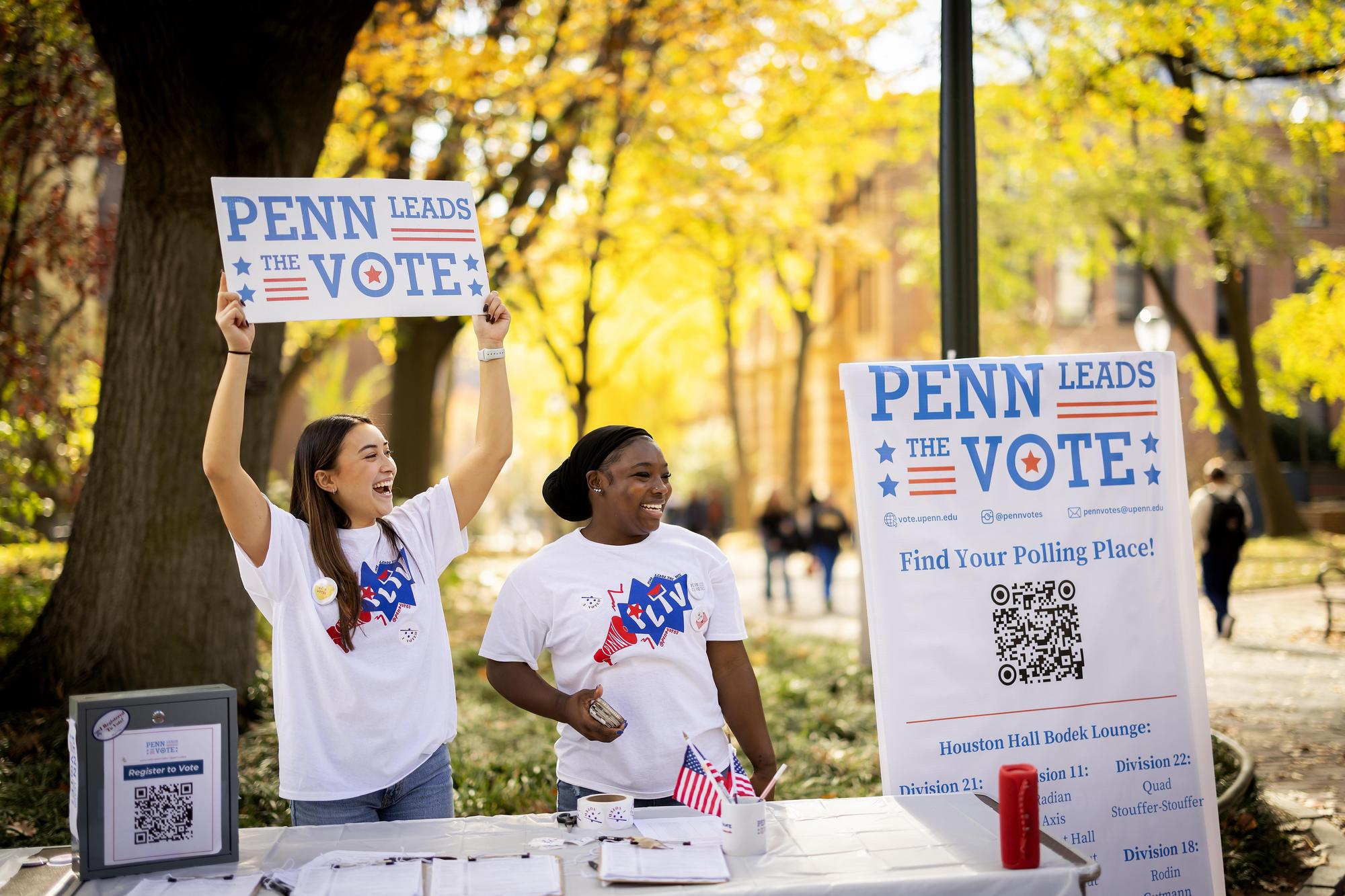 Students stand behind a Penn Leads the Vote table cheering on voters and holding a sign promoting voting.