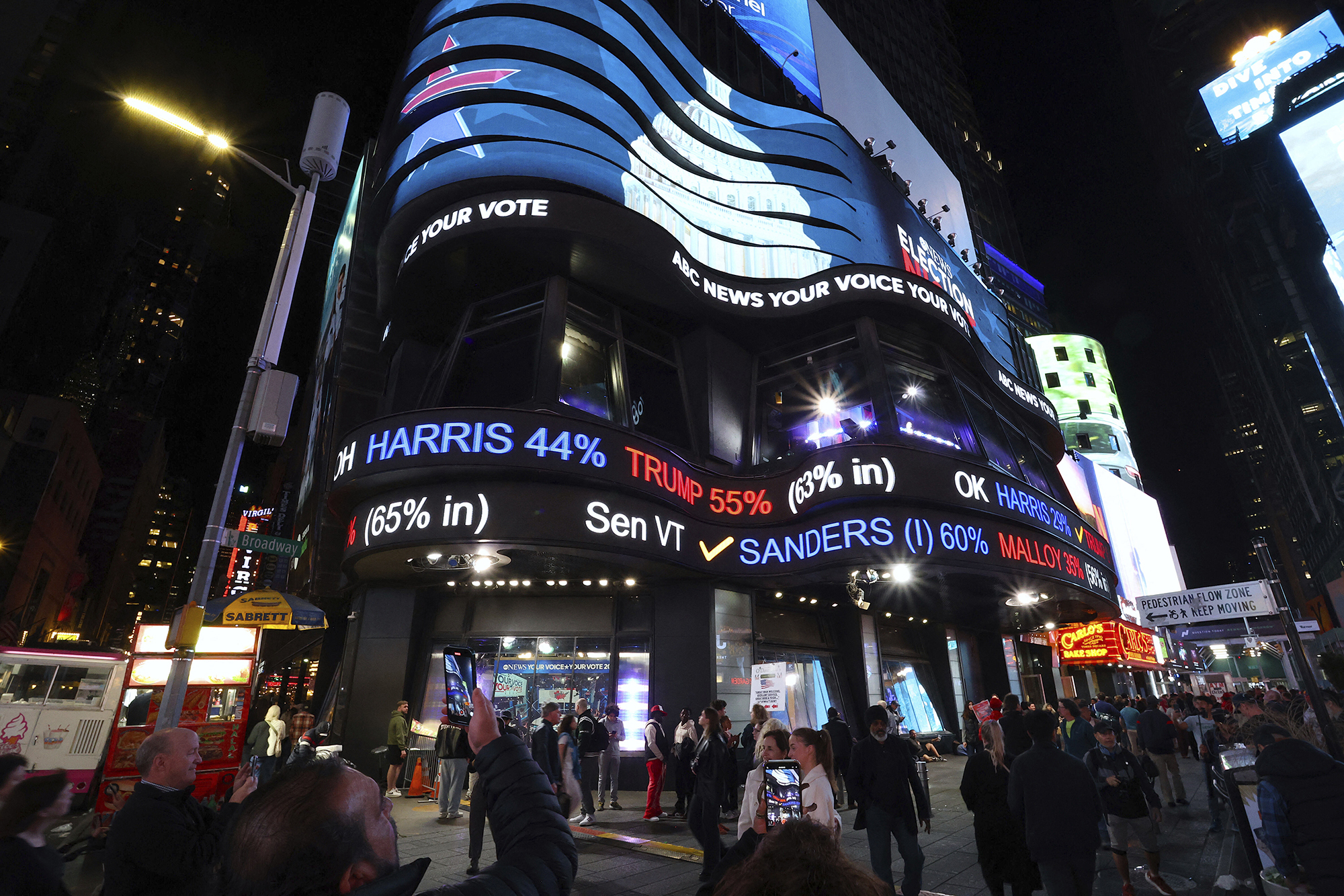 A side-scrolling election feed is displayed on the side of a building in Times Square in New York City. There are people below in the foreground.