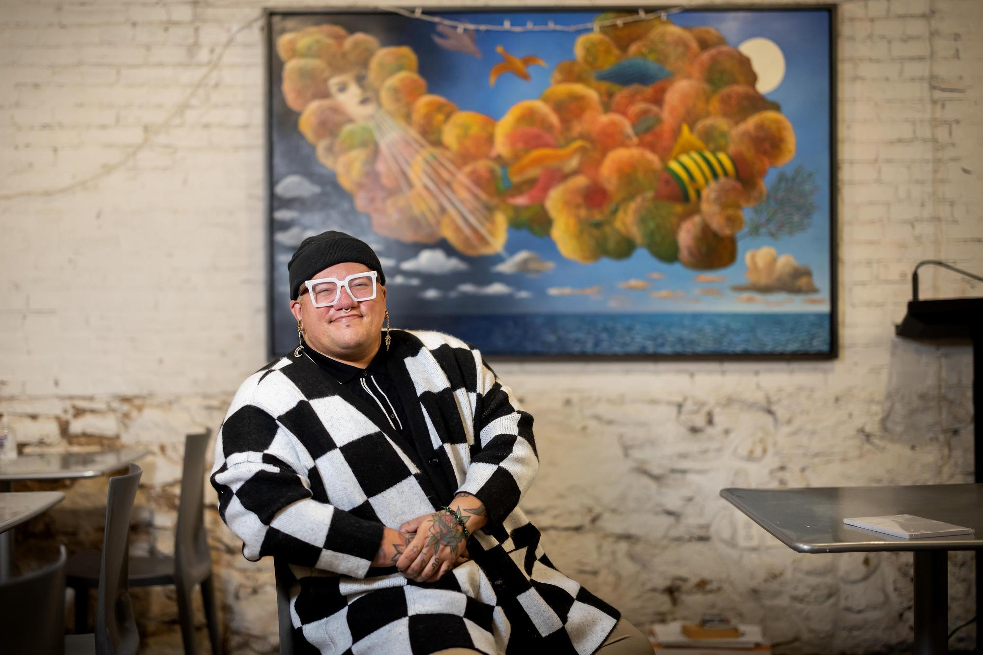 Eric Anglero sits at a table in the LGBT Center. They are wearing a black and white checkered sweater, white glasses, and a black beanie. 