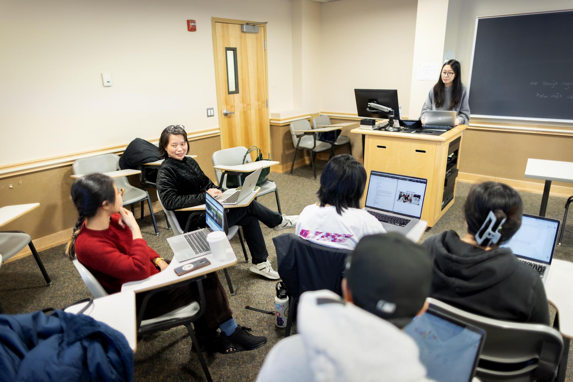 A classroom of students sitting behind desks. One student is at the front of the classroom delivering a presentation.