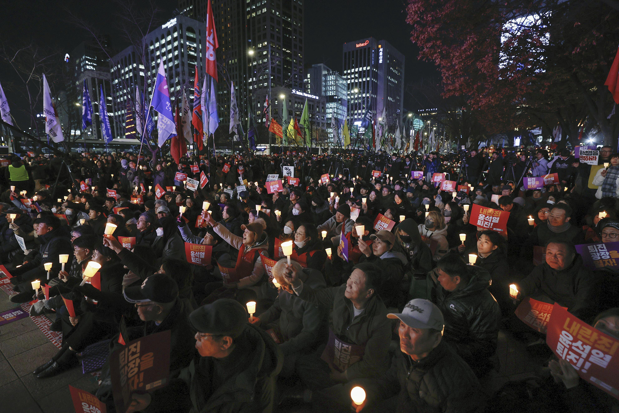 A crowd of people are gathered in a public square, facing left, holding candles and carrying signs in a vigil calling for the South Korean president’s resignation.