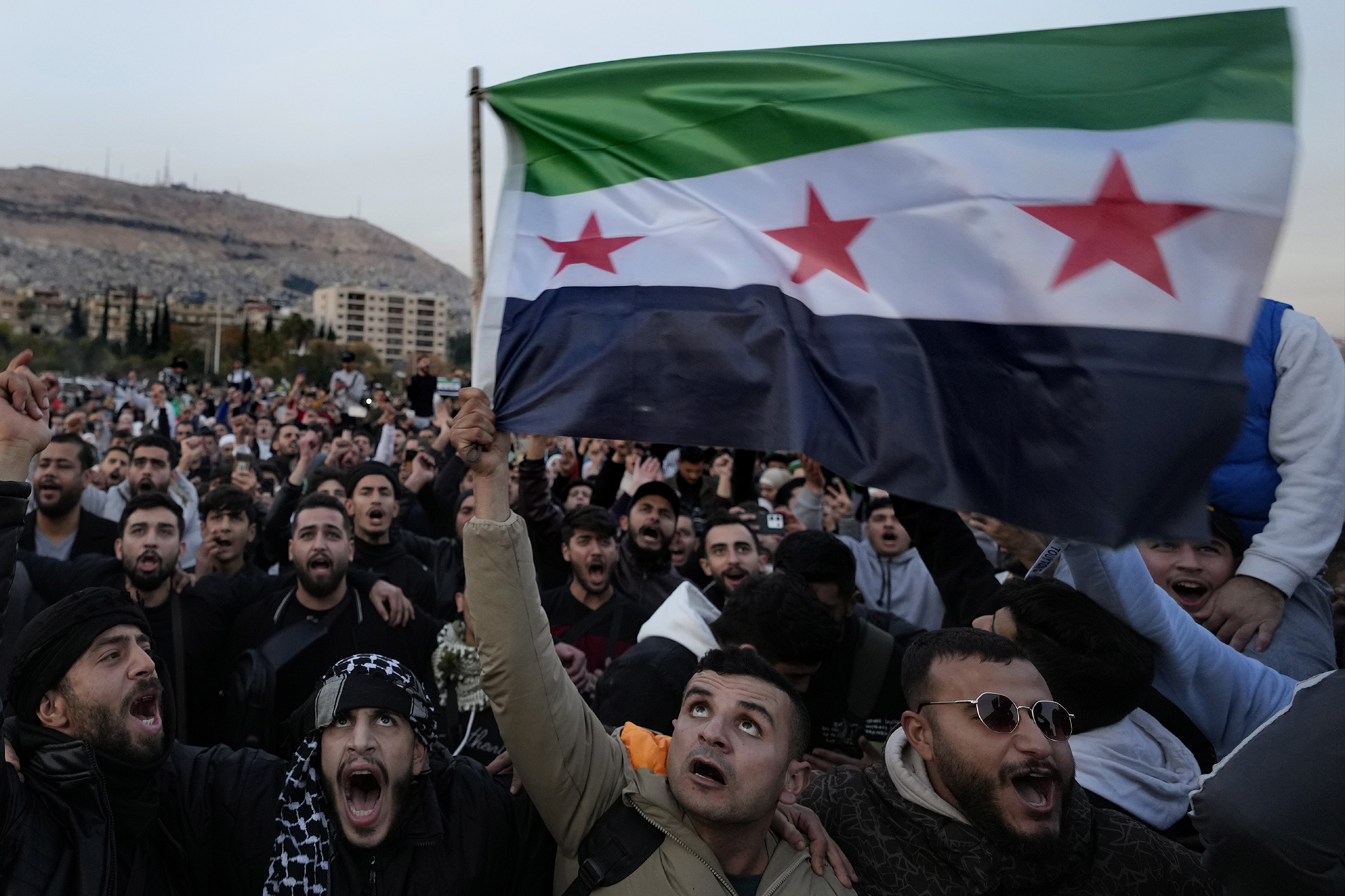 A crowd of Syrians gather in an open space. To the right, they are waving a revolutionary flag.