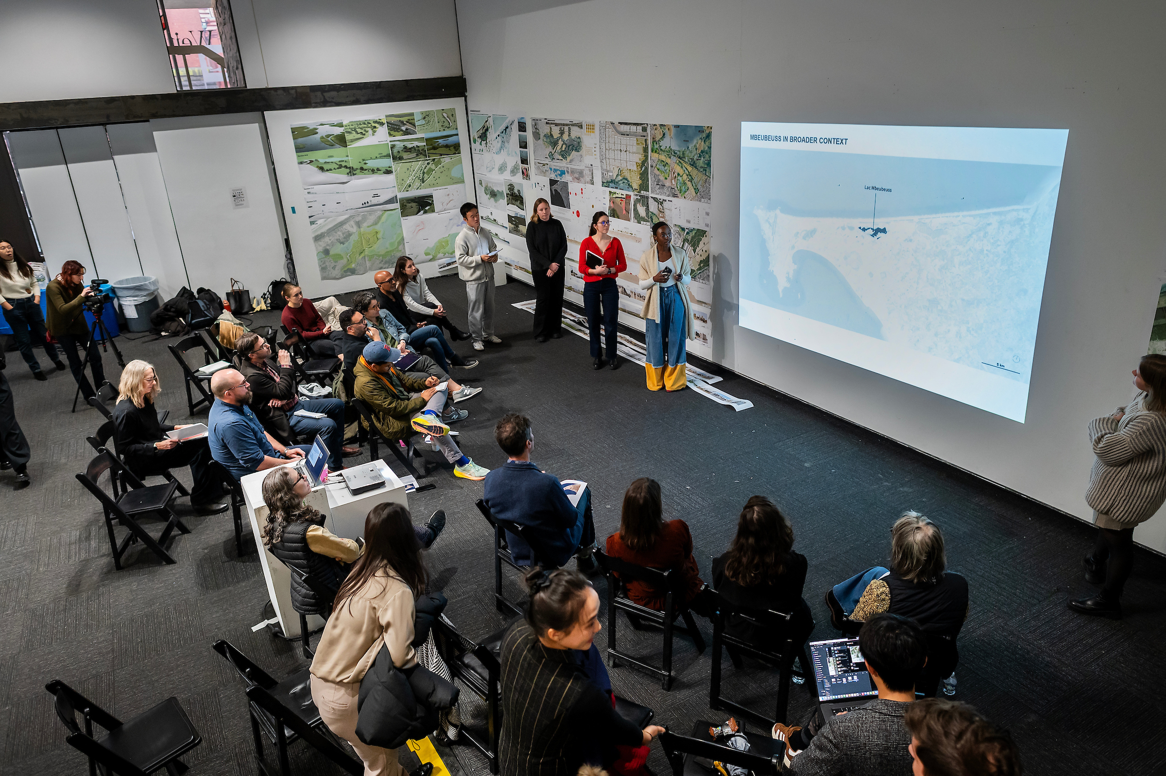 A group of presenters stands in front of a projected slide and large maps .