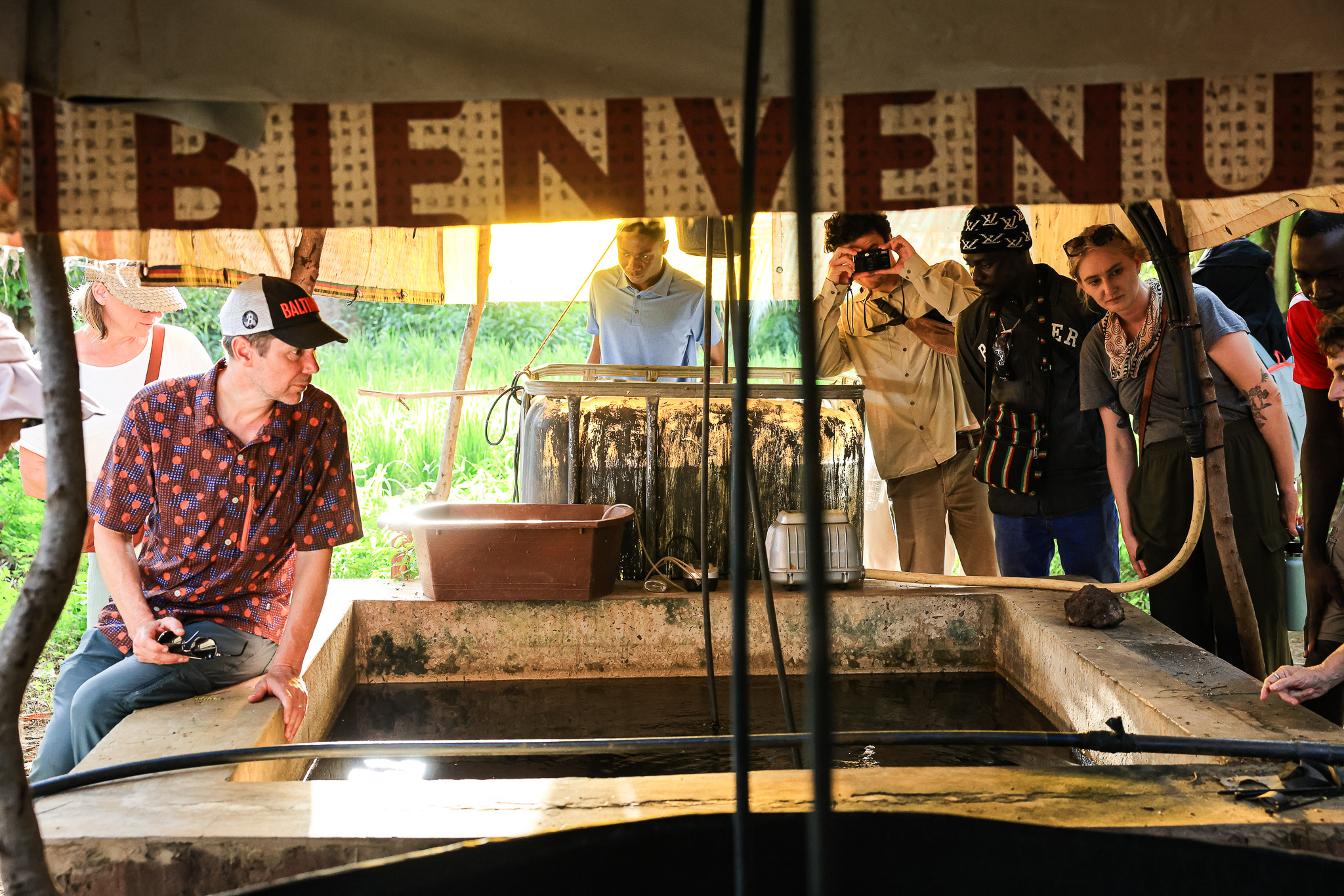 A group of people gathered under a shaded structure with a "BIENVENUE" sign.