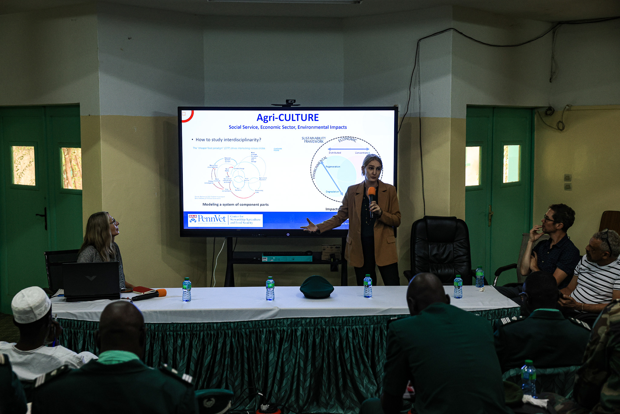 A presenter gestures toward a slide while addressing an audience in a classroom setting.