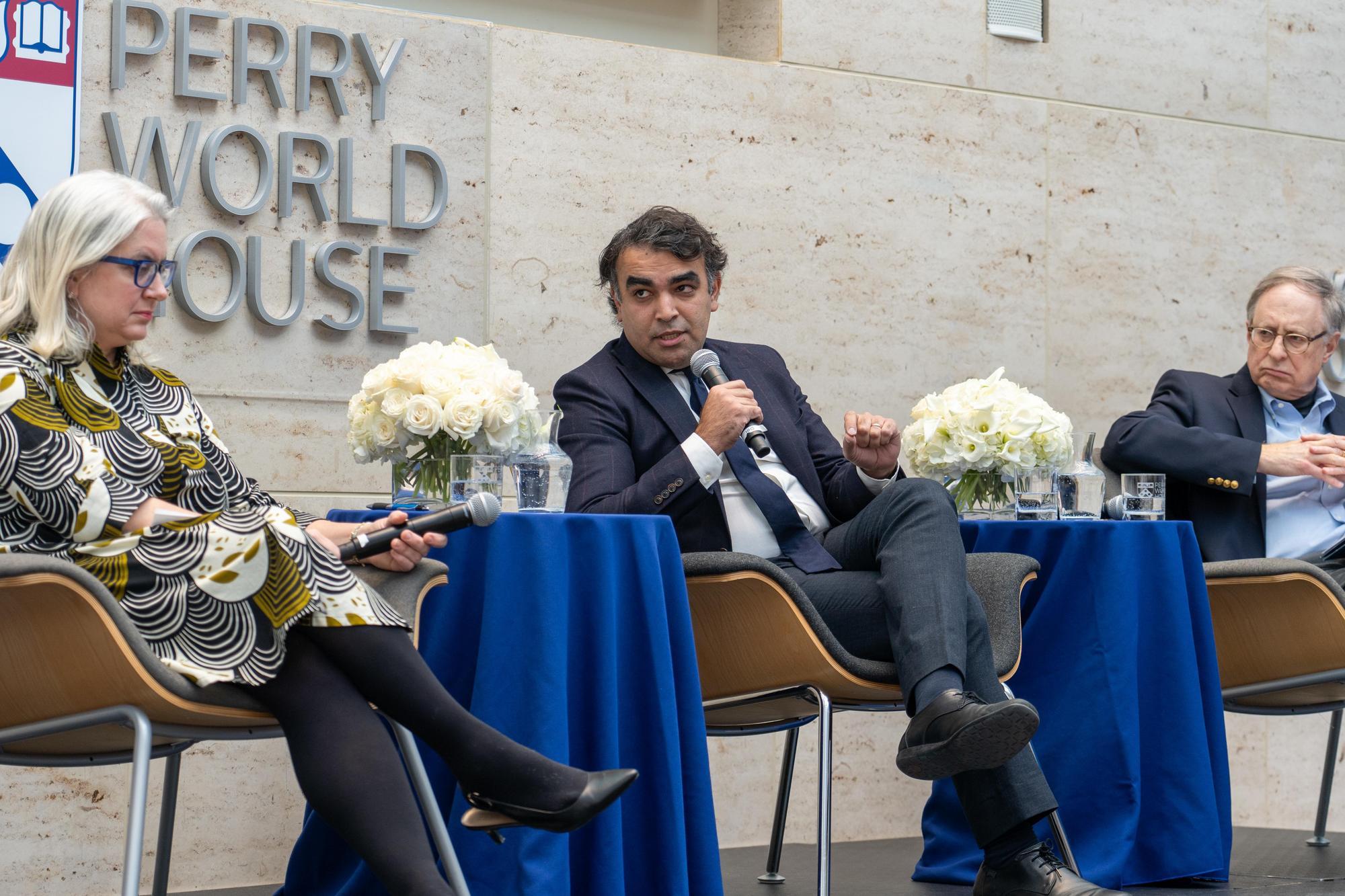 Three people sit in chairs on a stage in front of a Perry World House logo. The person in the middle is talking with a hand-held microphone.