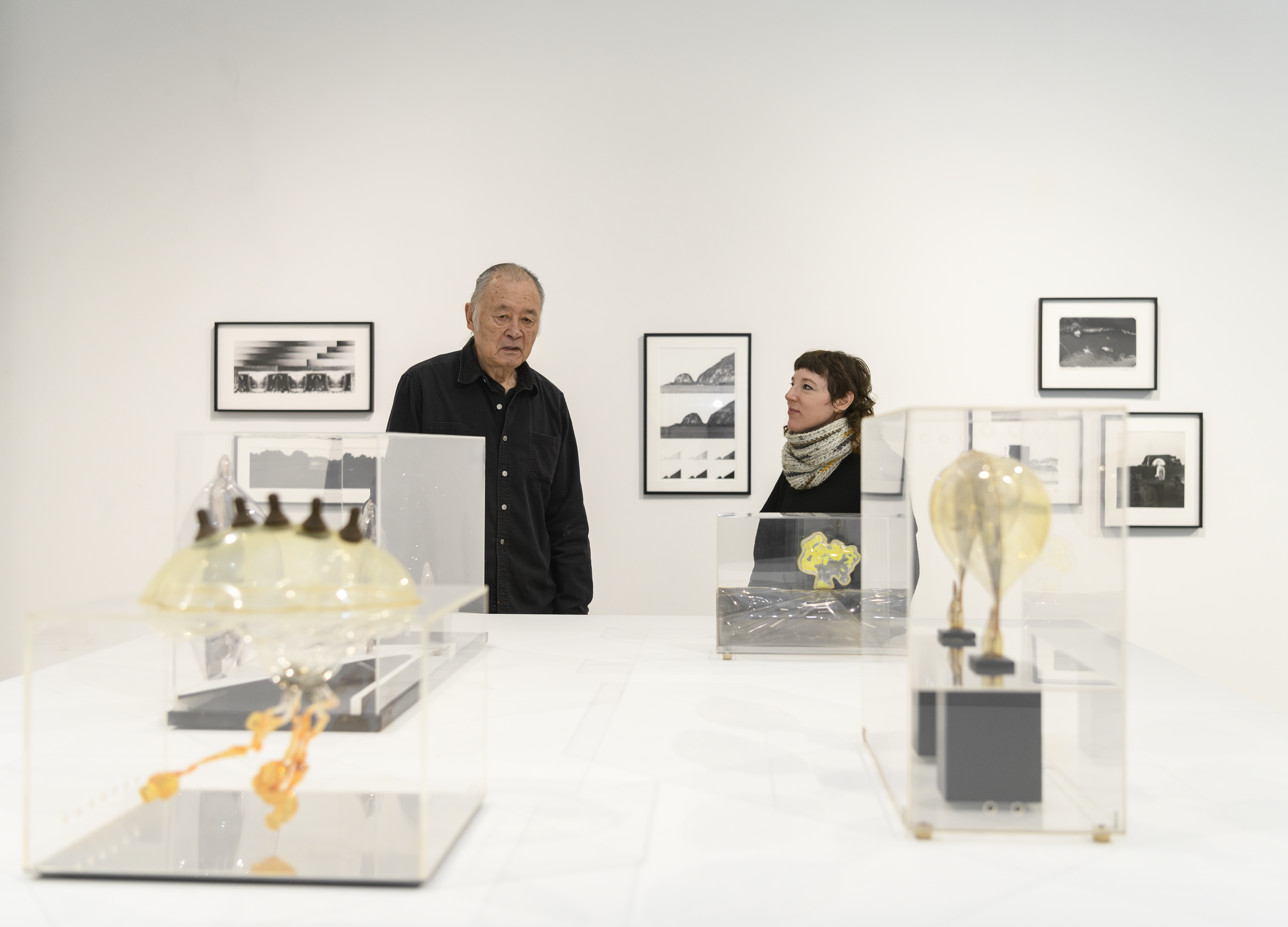 Artist Carl Cheng and curator Alex Klein in art gallery with sculptures on a table and photographs behind them.