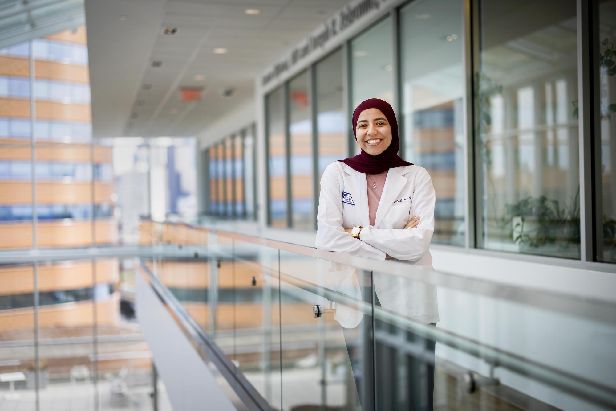 A smiling woman stands facing the camera with her arms crossed. She is standing in front of a glass-fronted row of offices with a glass railing in front of her. She is wearing a white coat and maroon head covering.