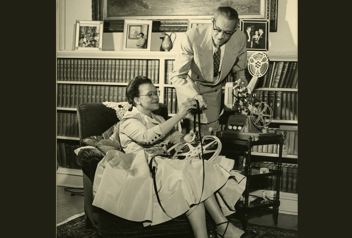 Sadie and Raymond Alexander with a film projector in a room with books on bookshelves and framed photos behind them. 