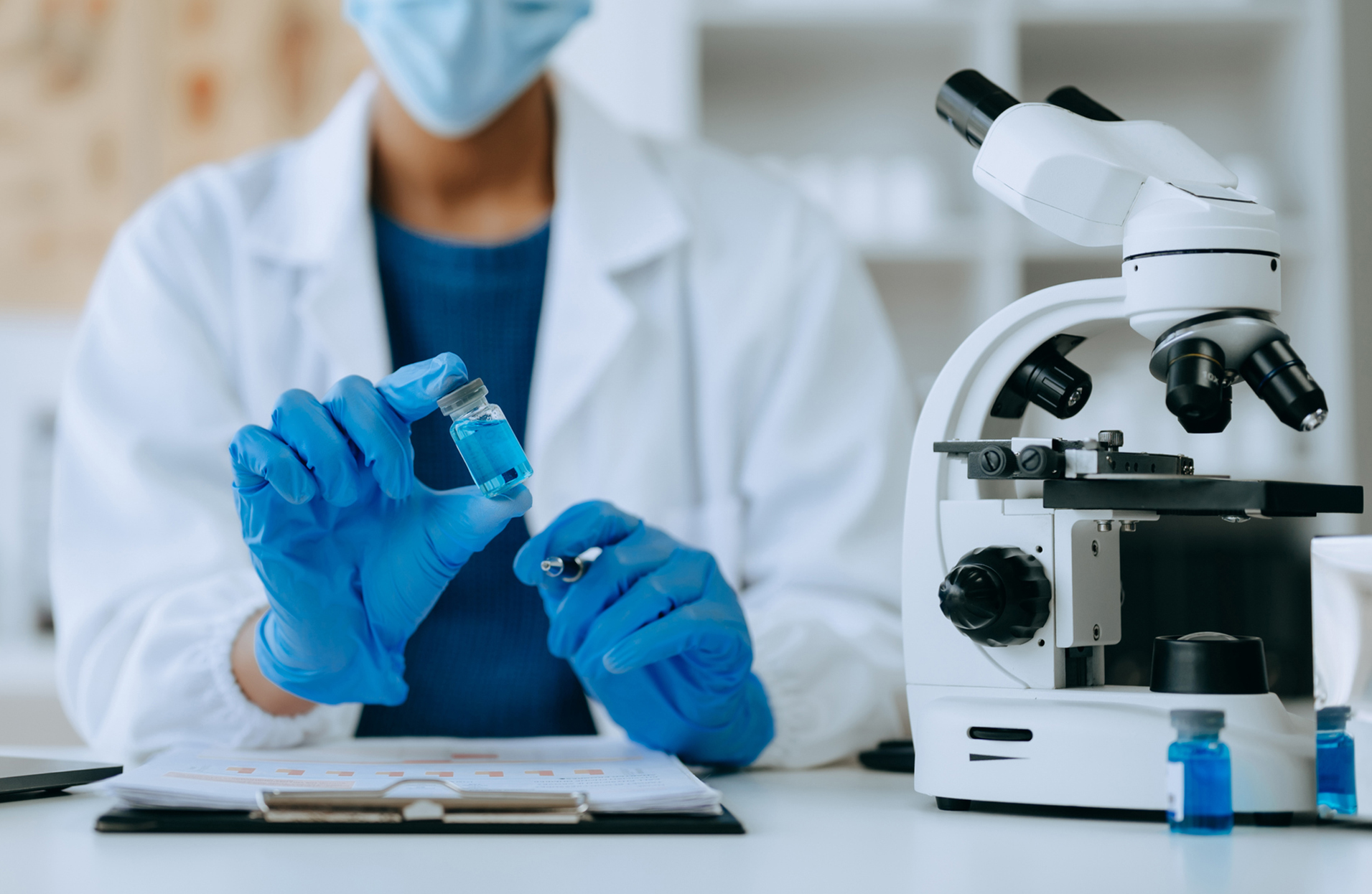 A researcher holding a vaccine vial in a lab next to a microscope.