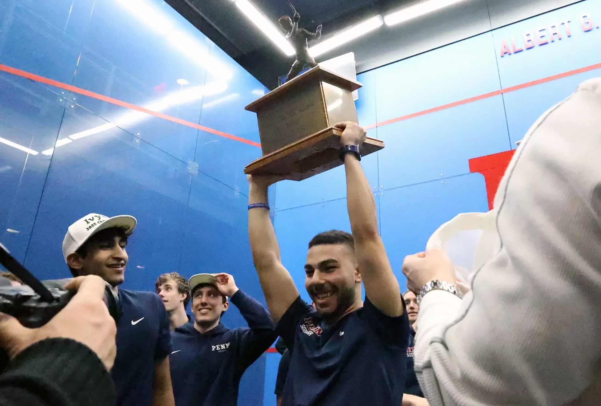 A member of Penn’s squash team holds a squash trophy over their head with other teammates.