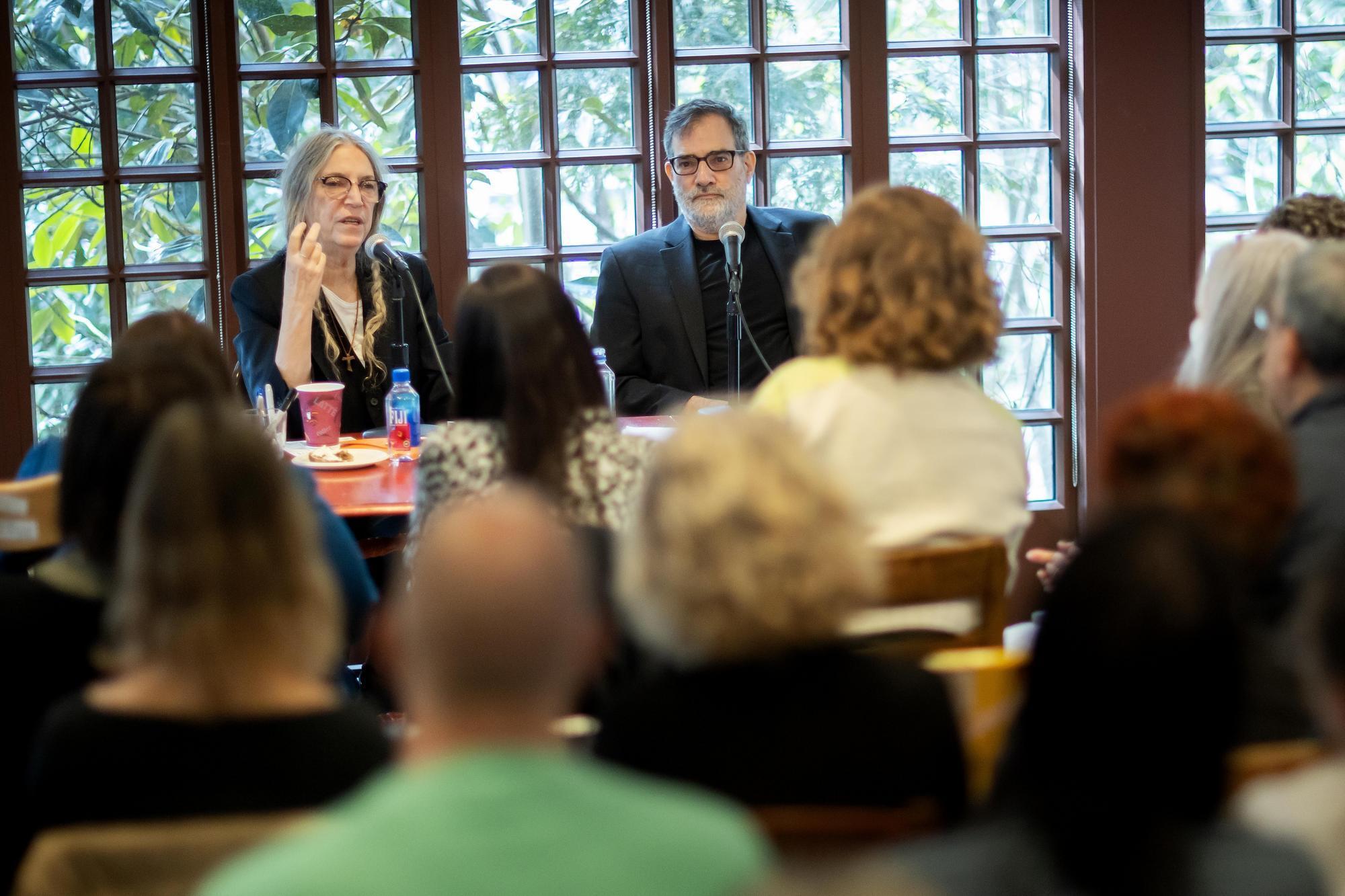 Patti Smith and Al Filreis sitting at a table with in a wall of windows behind them and an audience in front of them