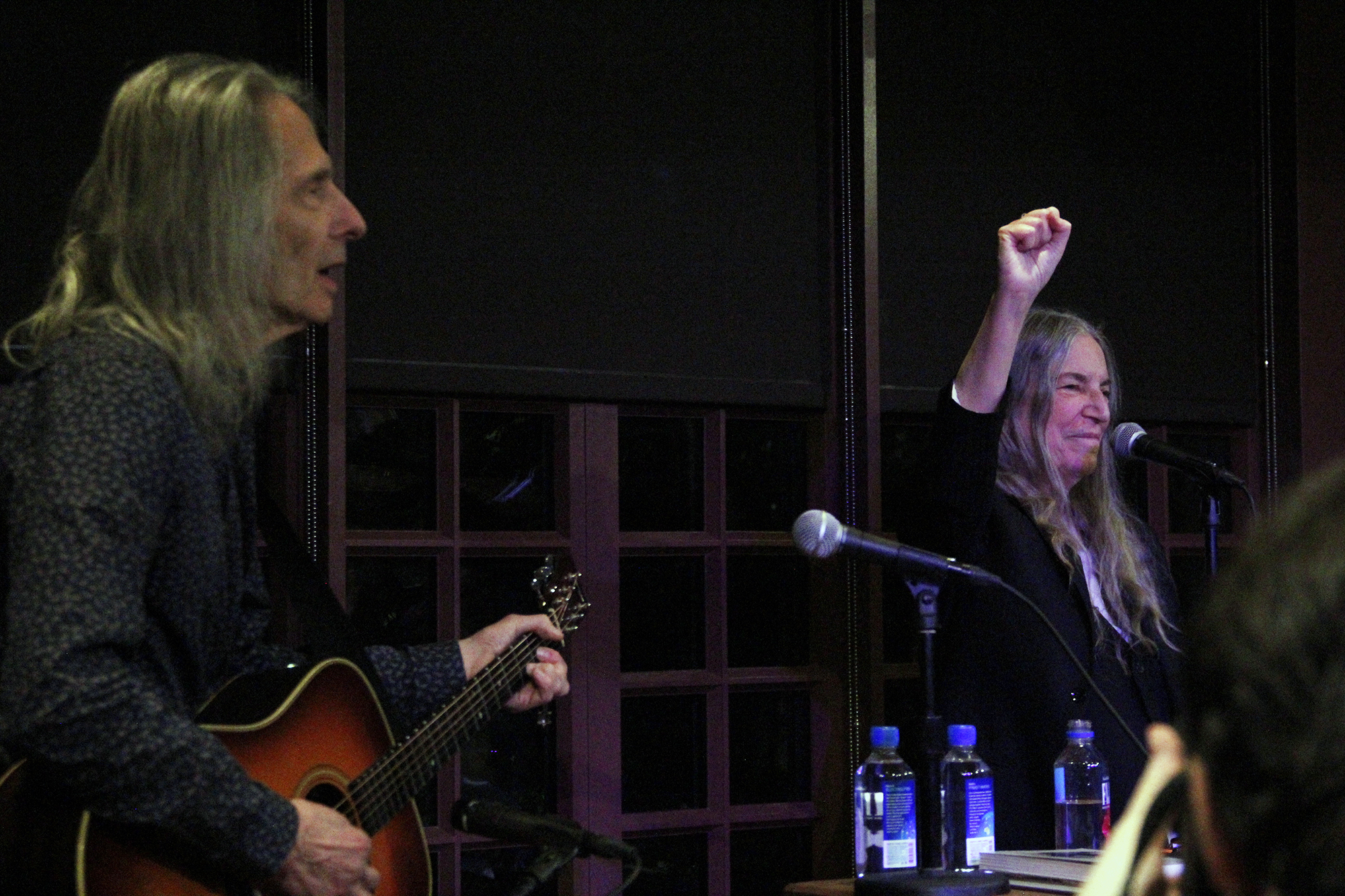 Patti Smith at a microphone holding her fist in the air and Lenny Kaye playing the guitar and singing next to her. 