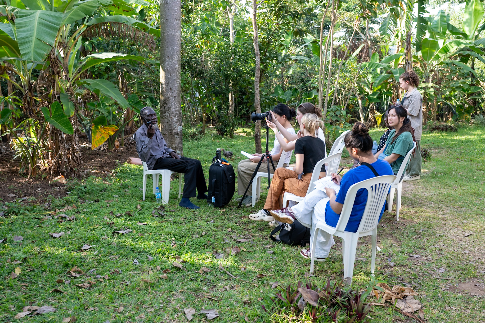 Several students sitting in chairs under a tree in a lush forest setting up a camera with a person in a chair facing the camera