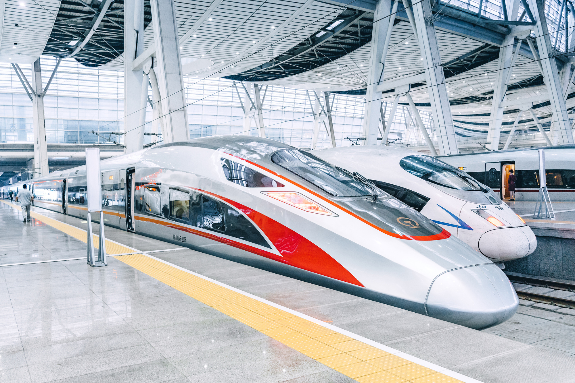 Two bullet trains sit side-by-side in a silver-and-white train station. The train closest to the camera has red markings on the side and top.