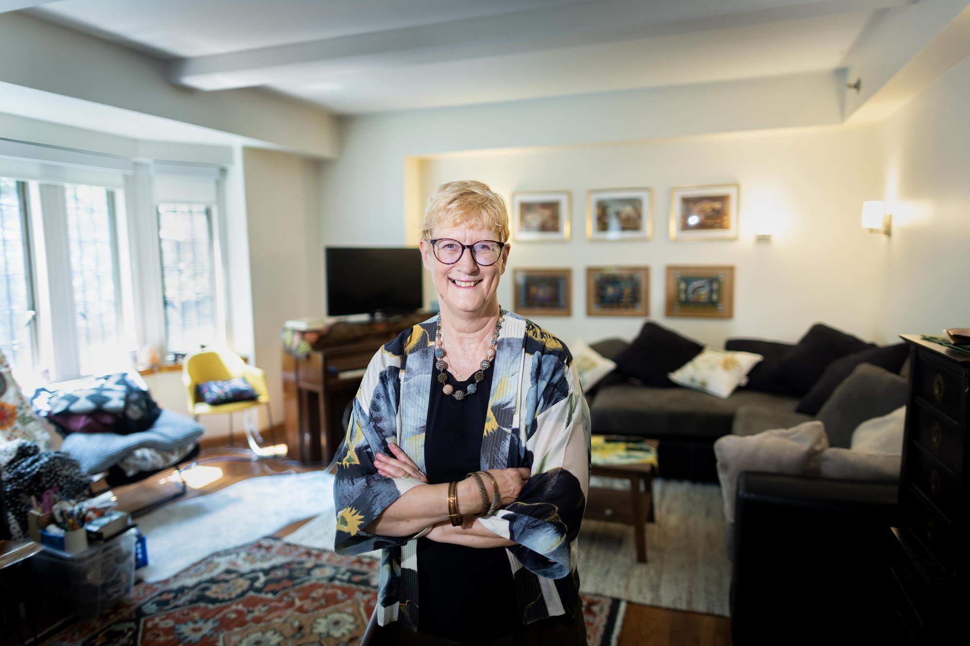 A smiling woman stands in front of her living room with arms folded.