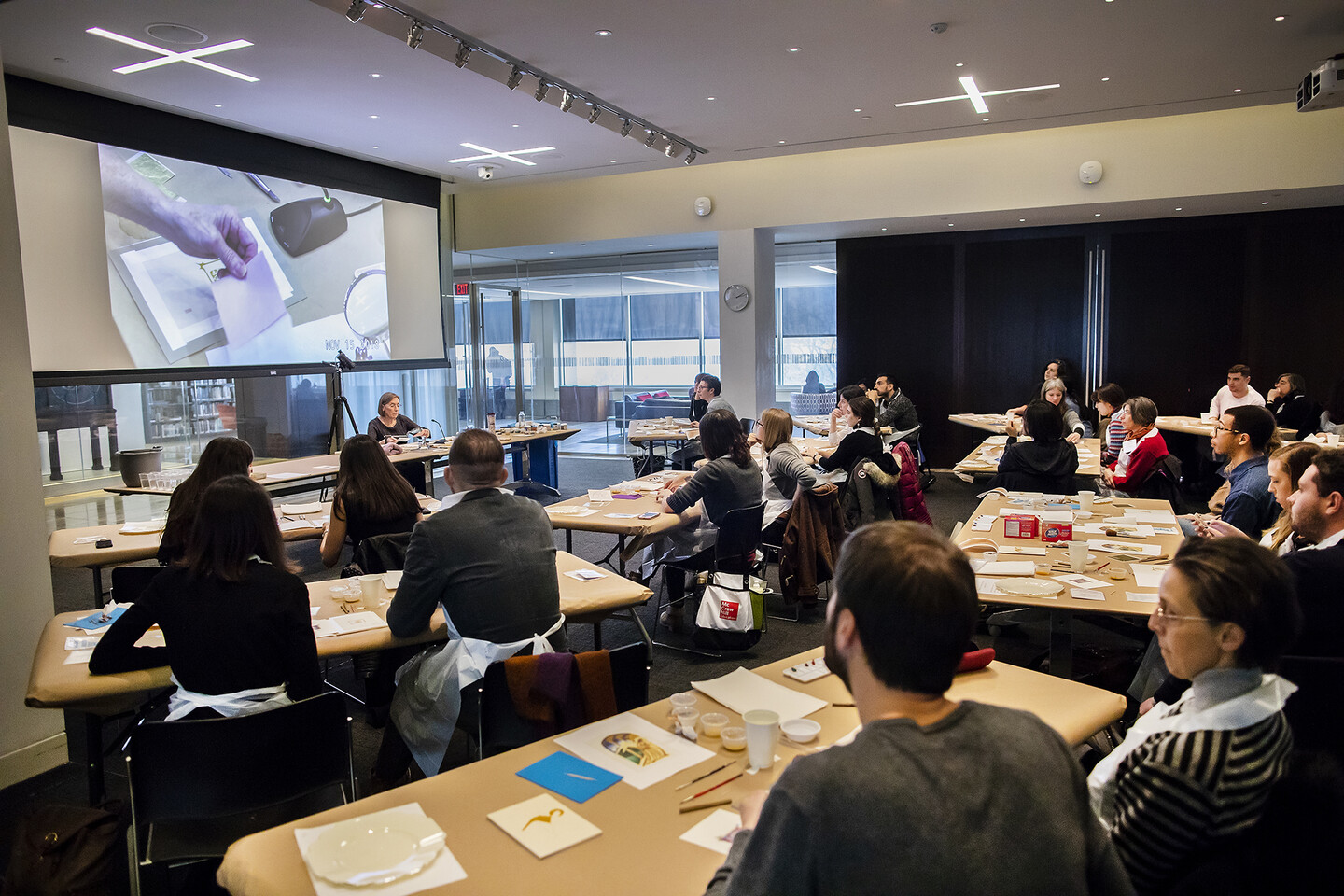 many people sitting at tables looking up a screen to see demonstration of working with materials to make illuminated letters