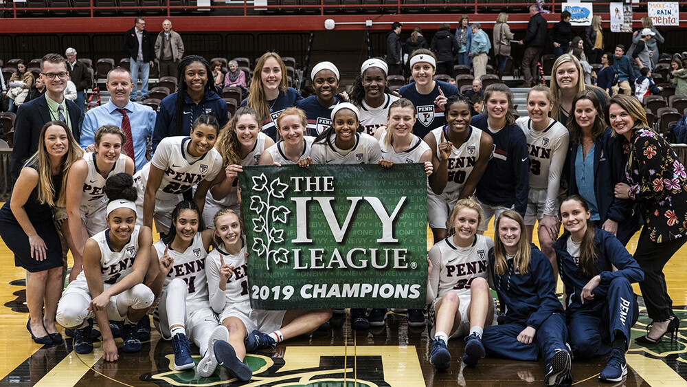 Members of the women's basketball team pose as a team with a banner that reads The Ivy League 2019 Champions on the court 