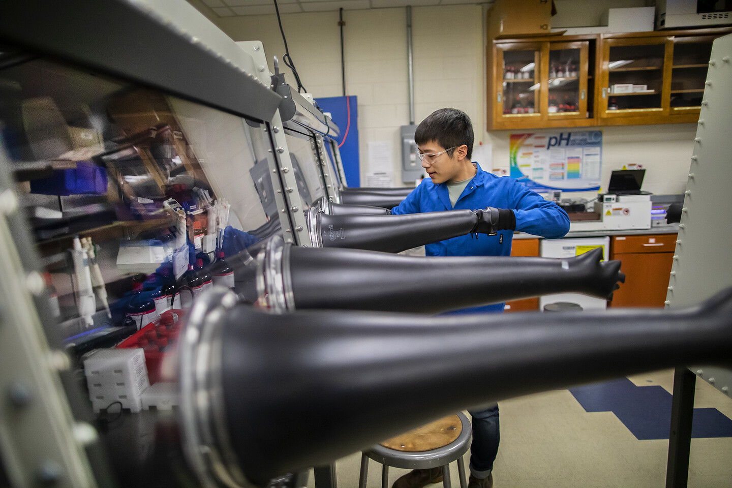a man putting his hand into a large glove that is sticking out from a glass box inside a chemistry lab