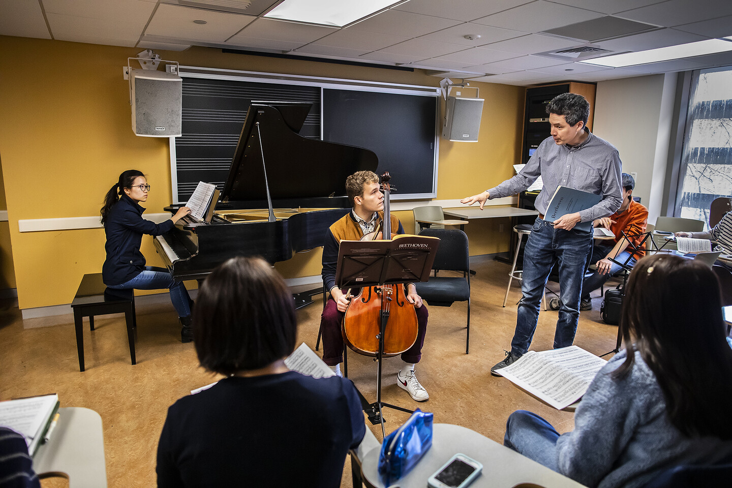 Students play piano and cello while instructor stands and speaks to them. 