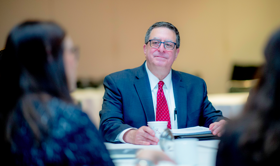 Person seated at conference in suit and red tie looks at another speaker in forground