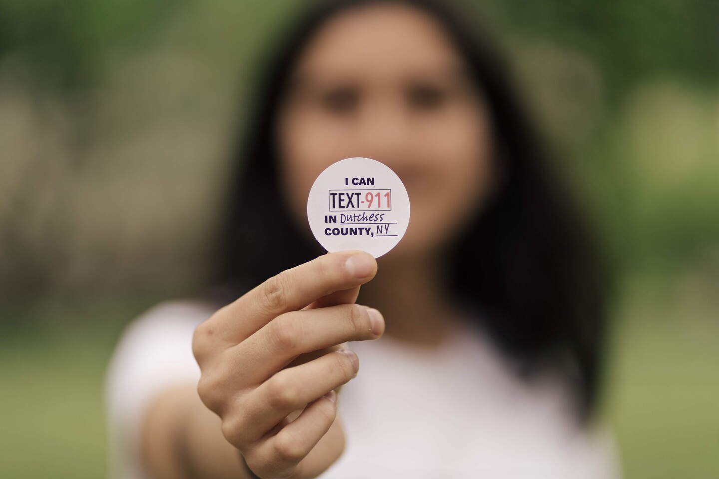 A blurry person holding a sticker that says "I can Text-911 in Dutchess County, NY"