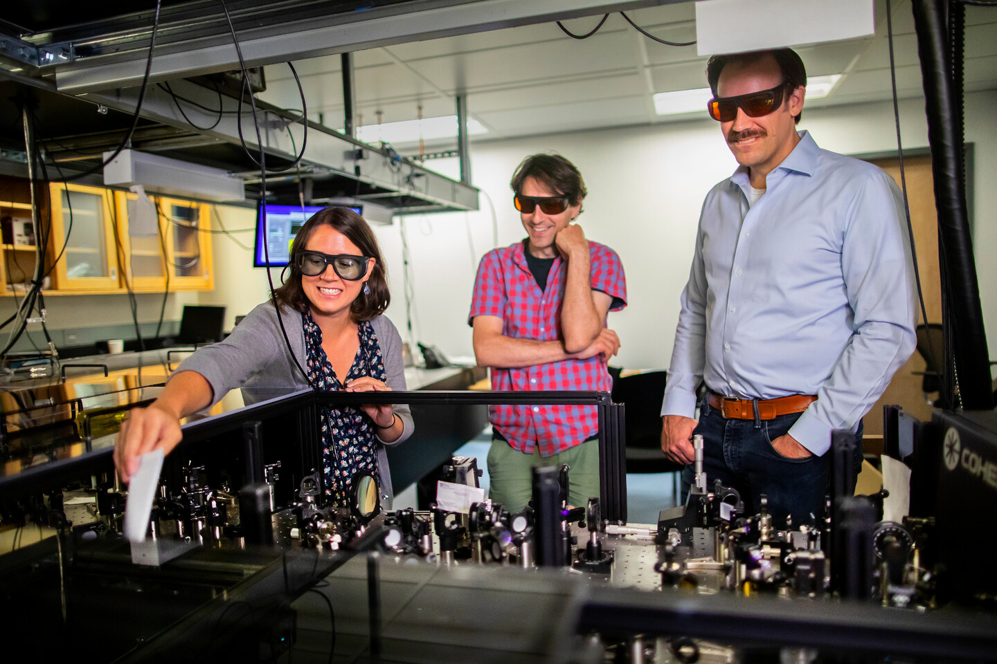 anna, subotnik, and schelter looking at an optics table, anna is holding a piece of paper to catch the laser light at the far end. 