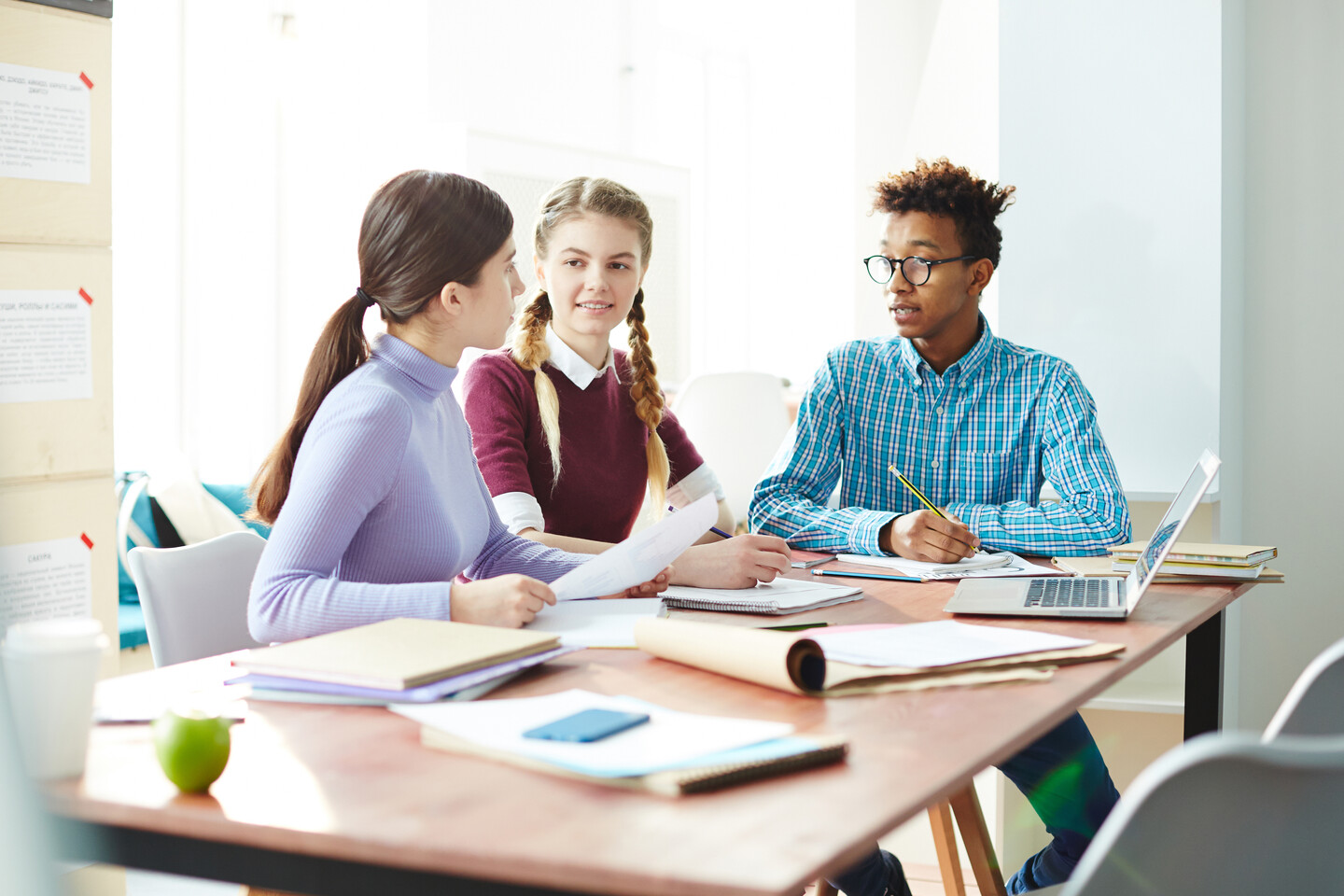 Three students engaged in conversation sitting at a desk covered with papers, notebooks, and a computer.