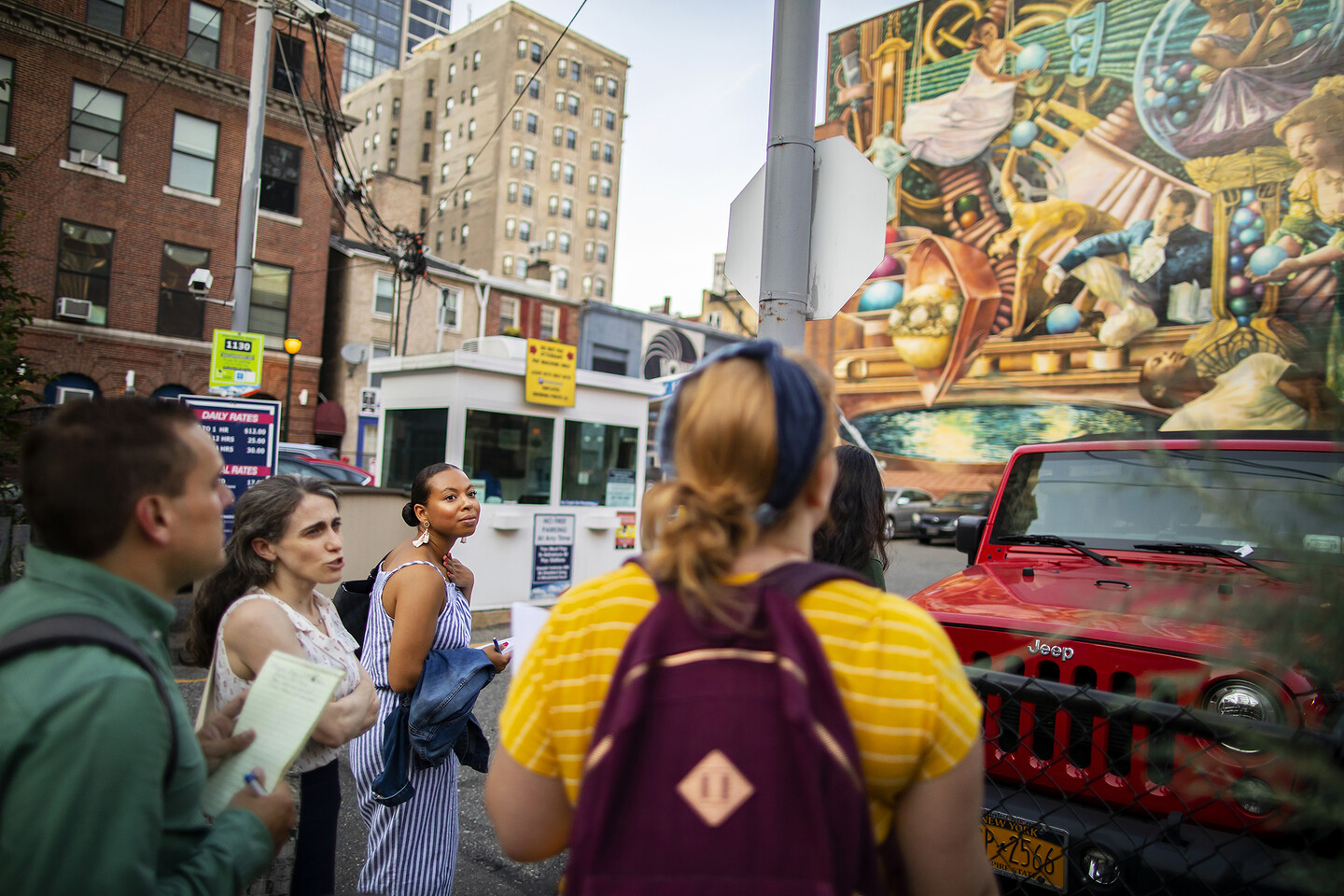 A group of people standing in front of parked cars, one speaking while the others listen and look at the mural on a building wall.
