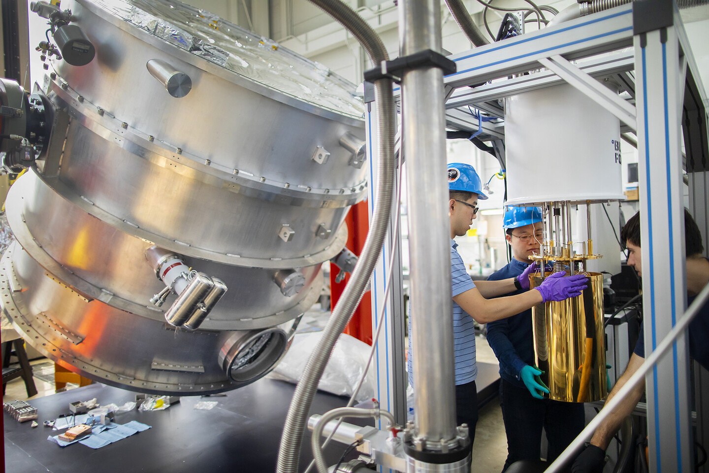 students holding a gold casing around metal tubing and wires, with a large metal cylinder in the background