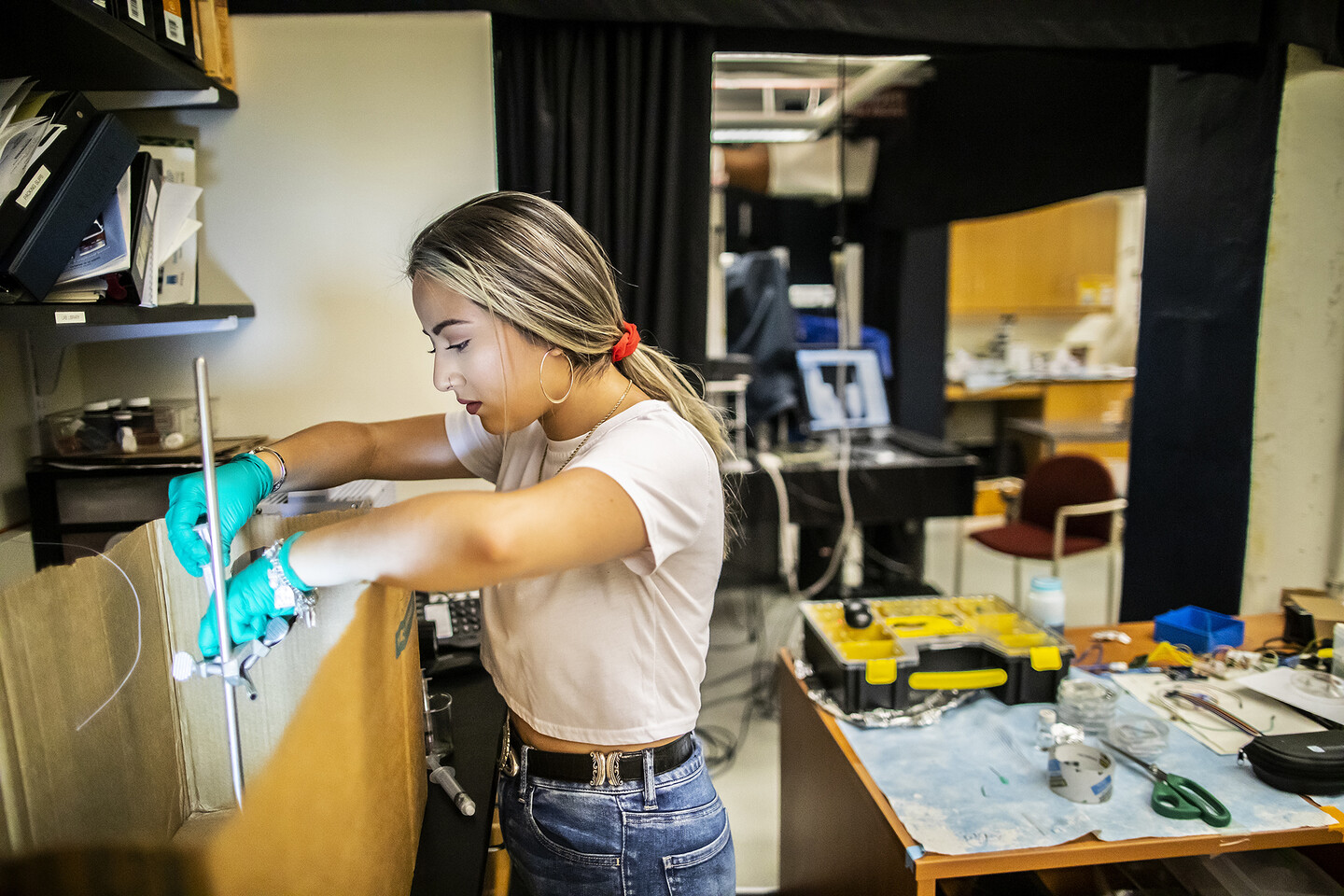 padilla adjusting a metal test tube holder that is held inside a cardboard box, working in a lab with tools and a computer in the background