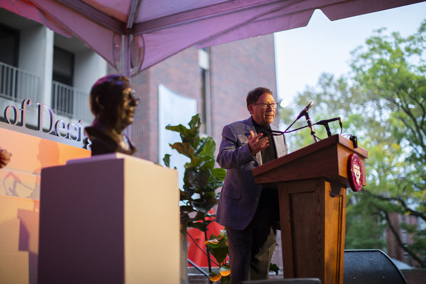 Stuart Weitzman talking at podium with bronze bust adjacent. 