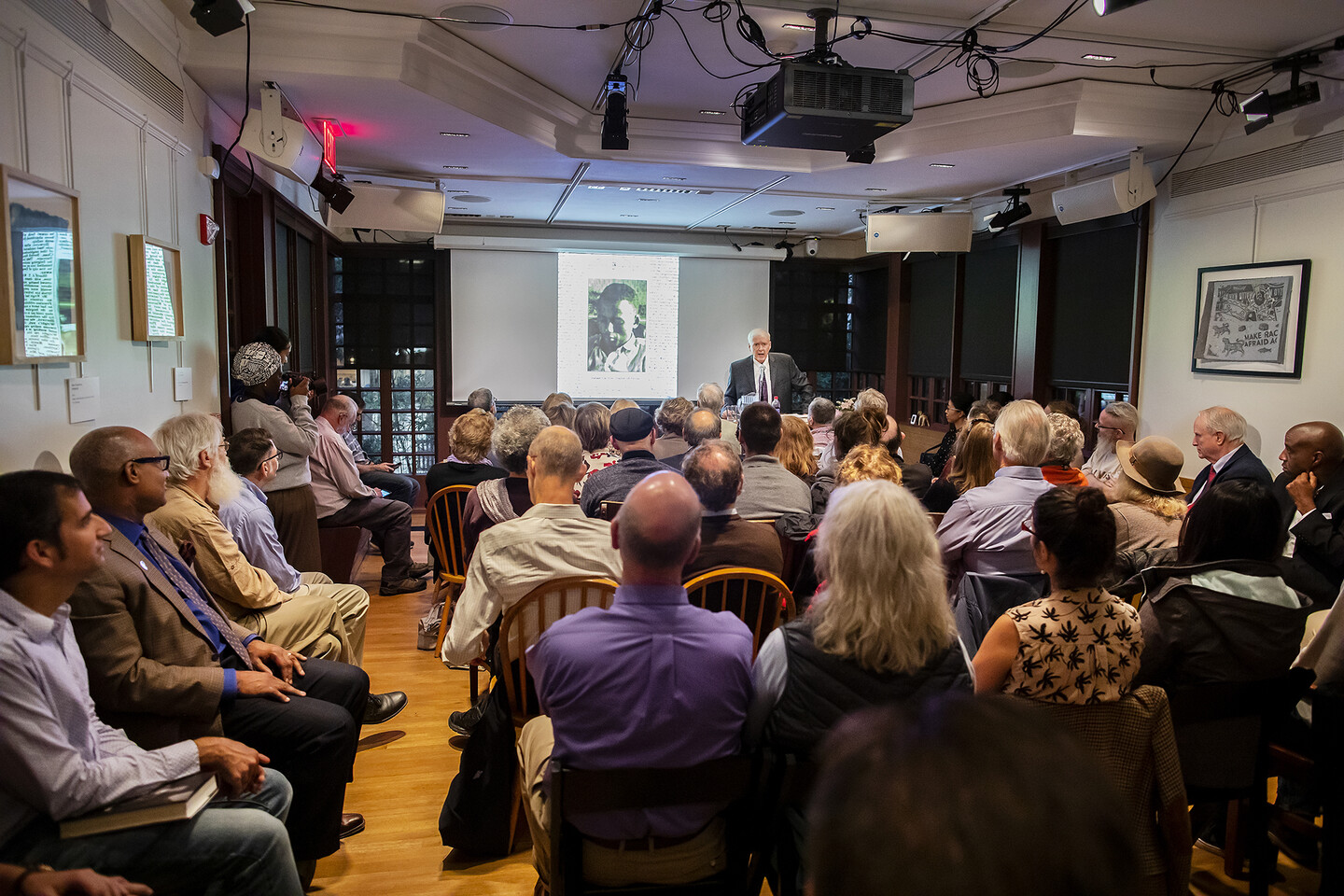 Paul Hendrickson speaking at a podium in front of a room filled with people seated and listening. 