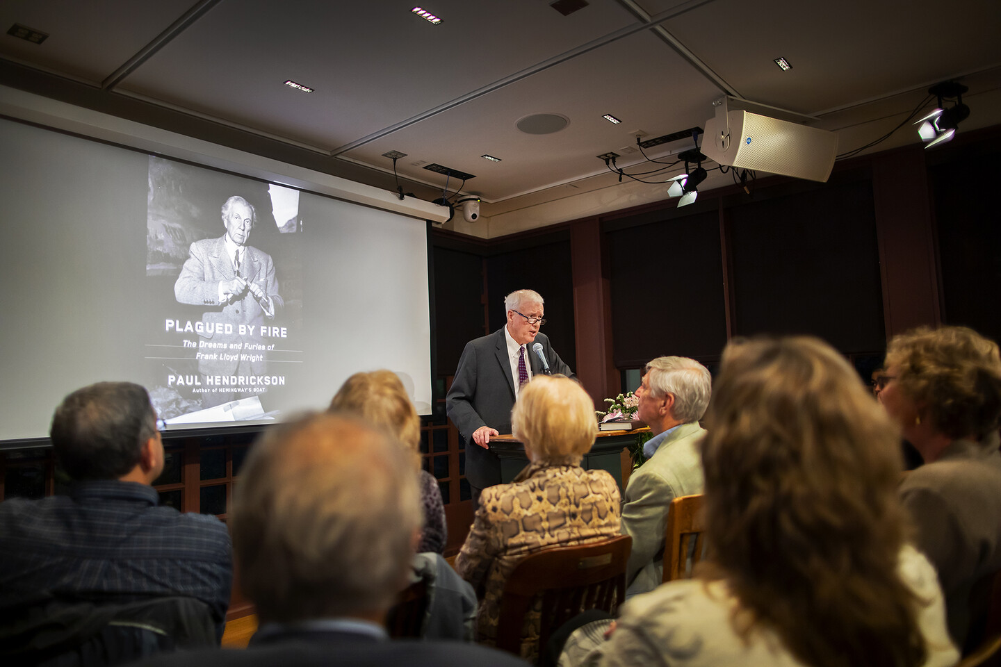 Paul Hendrickson speaking at a podium in front of a seated crowd with a photo of his book cover behind him. 