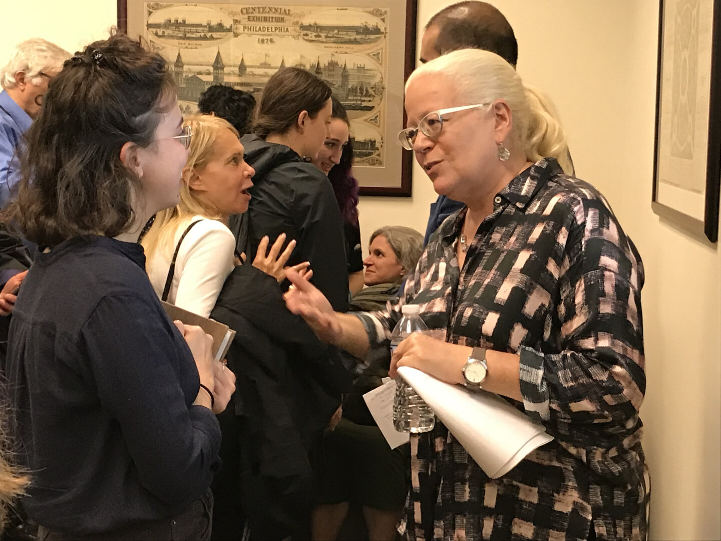 Two people facing each other, one whose back is to the camera. The other is holding a water bottle and a folded up piece of paper. They are in a crowded room.