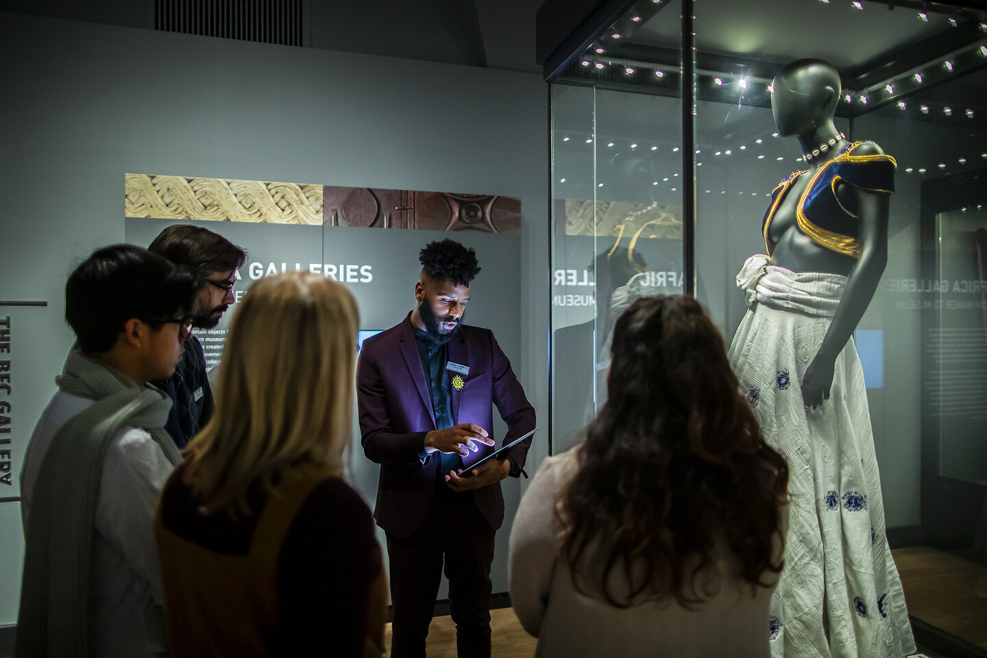 Man holding iPad addresses a group of four people beside a two-piece garment in a glass display case.