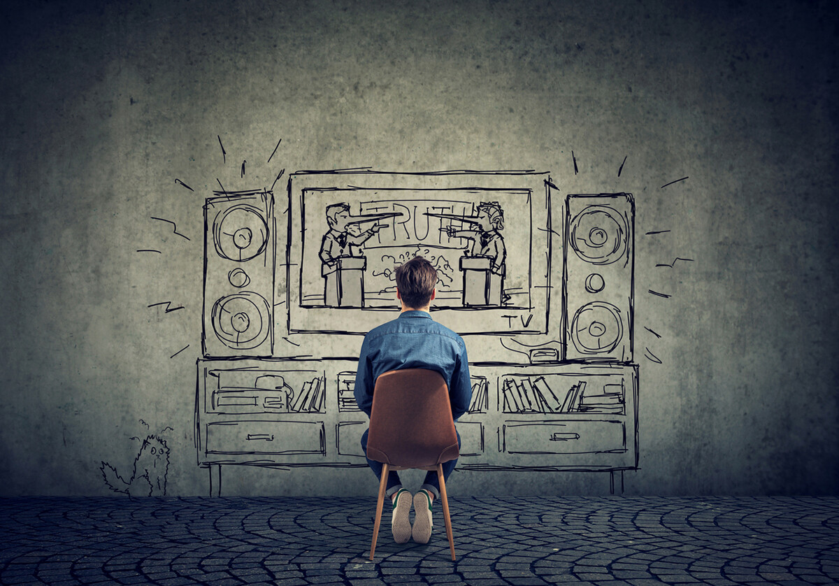 person in sparse room sits in chair facing a wall with a cartoon drawing of a televised political debate