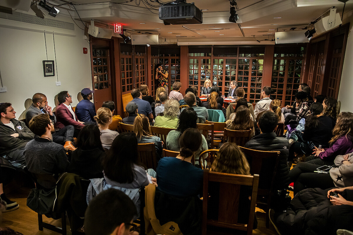 A group of people sitting in wooden chairs in front of a speaker who is sitting at a table at the front of the room.
