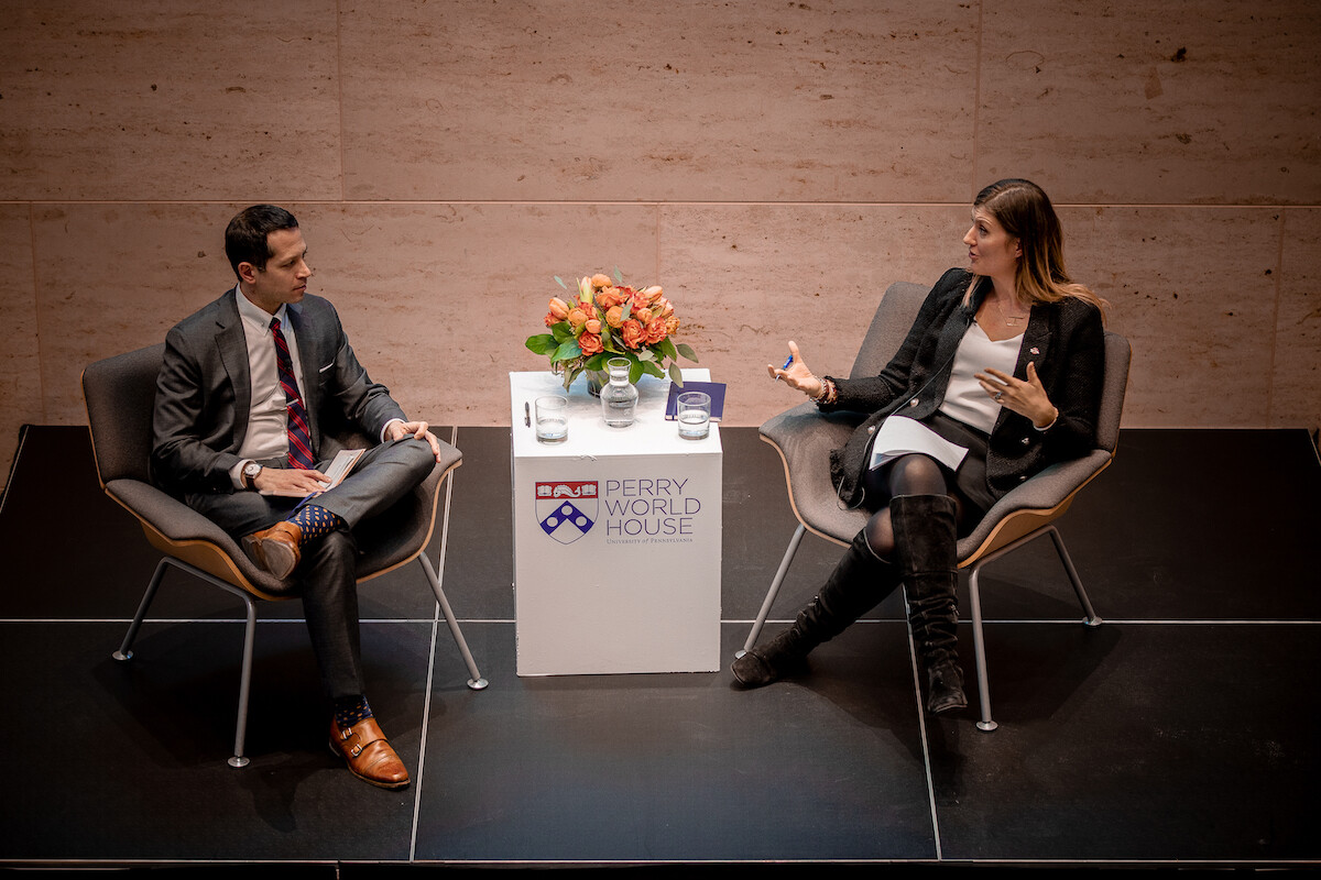 Two people sit in chairs in a discussion, with a table between them bearing a sign reading Perry World House, with a vase of flowers and glasses of water on top