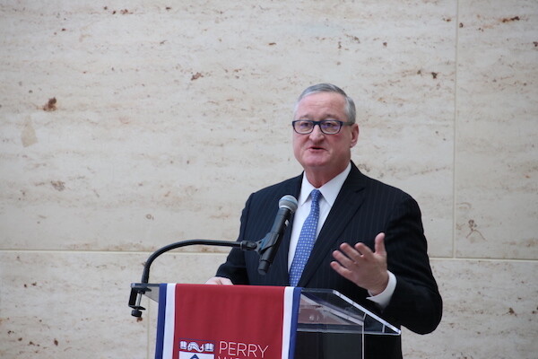 Person in black suit and blue tie speaks at podium with red banner, gesturing with left hand