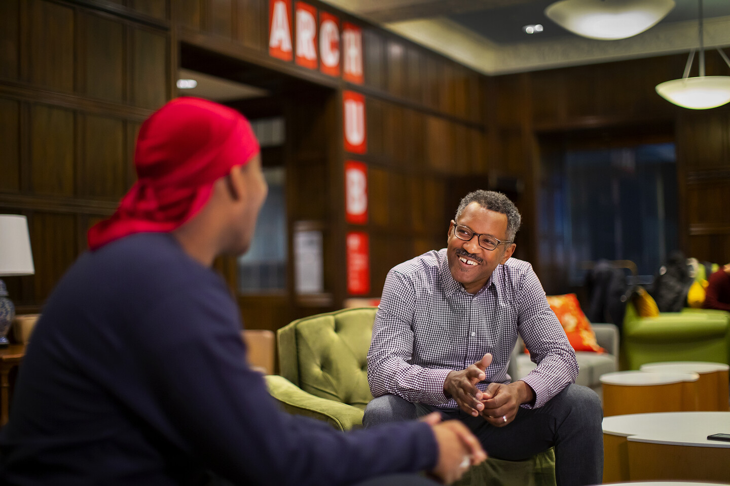 Two men talking and laughing in a student lounge. In the wood paneling in the background, a sign reads "ARCH" 