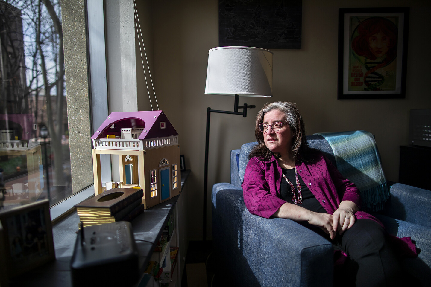 woman in an armchair looks out a window; a dollhouse and books are on the windowsill