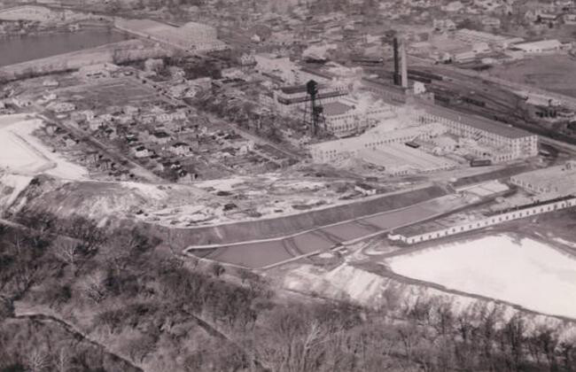 Historic aerial view of a parcel of land containing two towers and densely packed industrial buildings adjacent to a town.