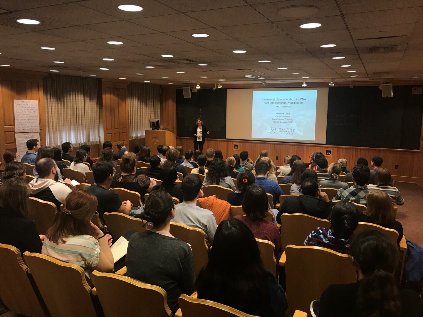 an audience listening to a presentation in a lecture hall by a speaker at the front of the room