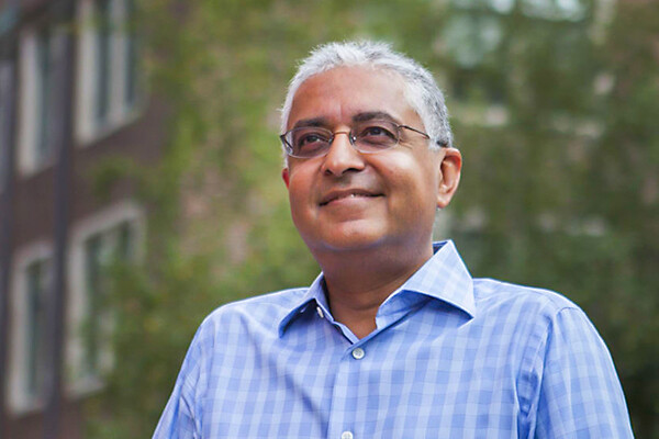 Person with glasses smiles in front of blurry trees and a red brick building.